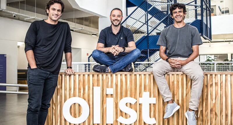 Líderes da Olist em frente ao logo da empresa, que anunciou a compra da Tiny ERP e Vnda Ecommerce